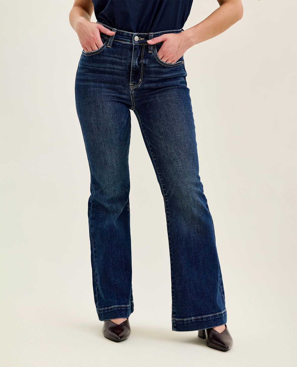 A person wearing dark blue high-rise flare jeans with hands in the pockets, paired with a navy top and dark brown pointed-toe heels, against a plain light beige studio background.