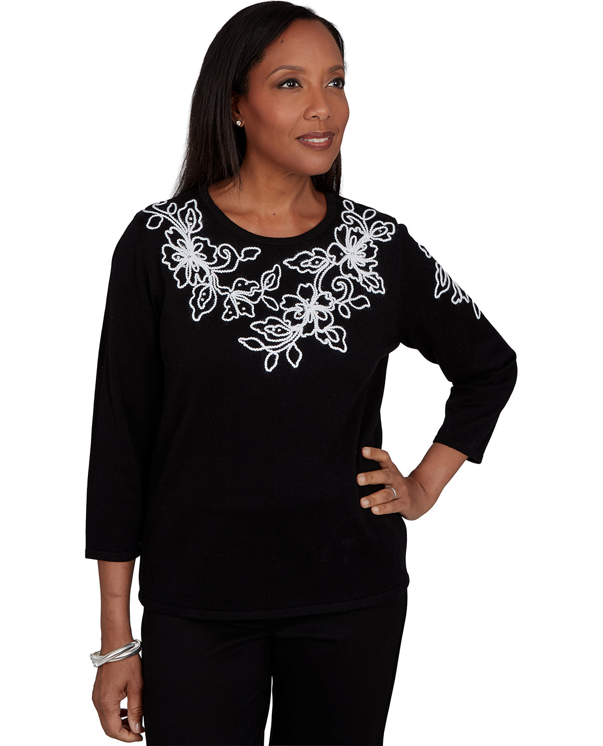 A woman modeling a black long-sleeve top with white floral embroidery across the chest and shoulders, paired with black pants, against a plain white background.