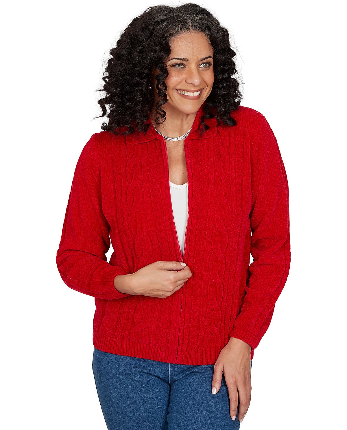 A smiling woman with curly dark hair wearing a bright red cable-knit zip-front cardigan over a white top and blue jeans, photographed against a plain white studio background.