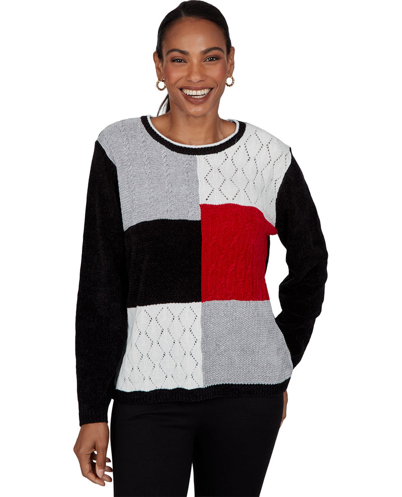 A woman wearing a color-block knit sweater with black, light gray, white, and red panels, paired with black pants, against a plain white background.