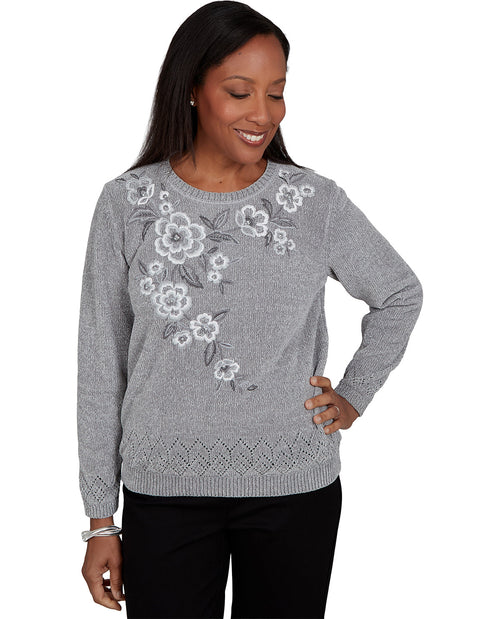 A woman wearing a gray knit sweater with white and gray floral embroidery on the chest, paired with black pants, against a plain white background.