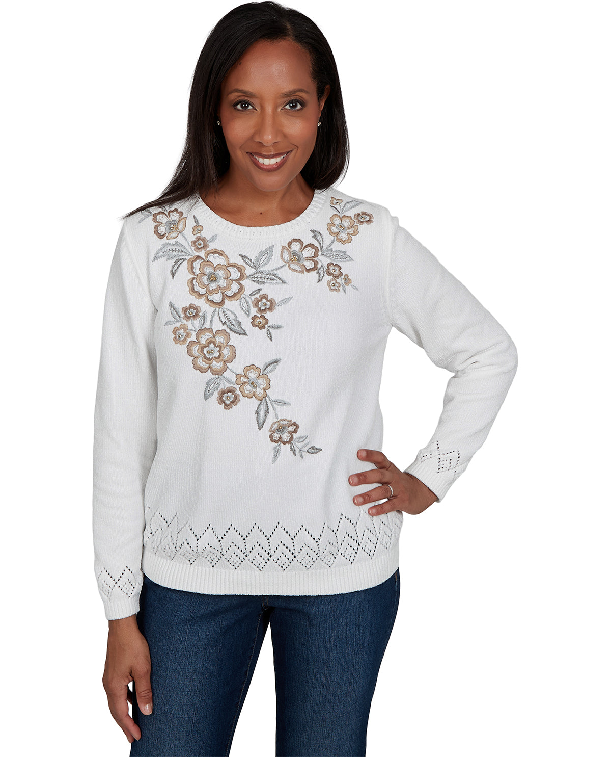 A woman modeling a white crewneck sweater with a floral pattern in beige and gray on the chest, paired with blue jeans against a plain white background.