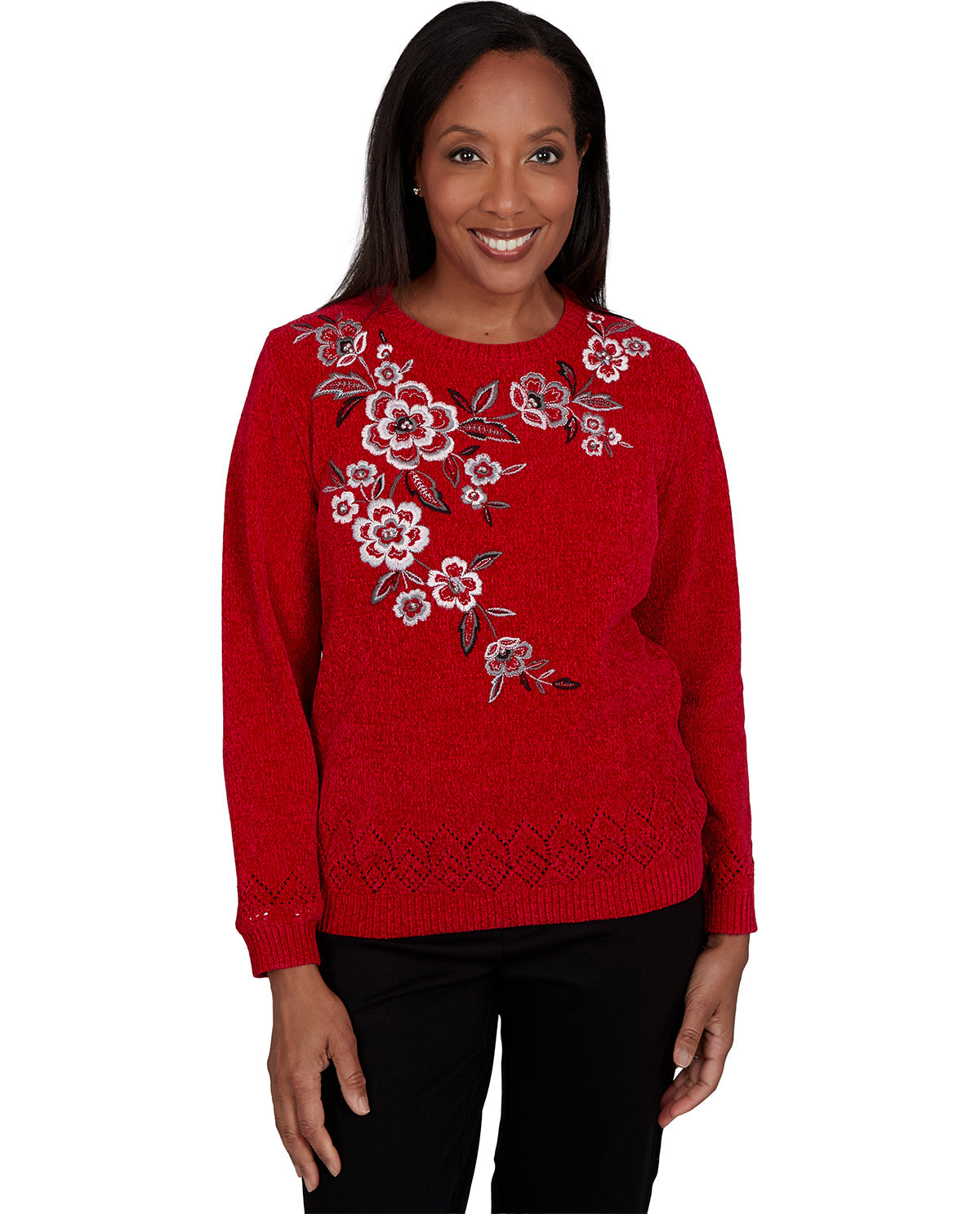 A smiling woman wearing a bright red knit sweater with white and pink floral embroidery and gray leaf details on the chest, paired with black pants, against a plain white background.