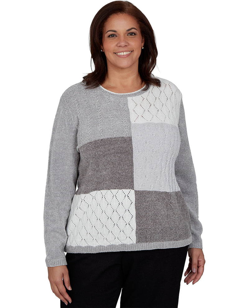 A woman wearing a gray and white patchwork knit sweater with black pants, photographed against a plain white background.