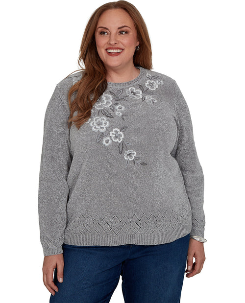 A woman wearing a heather gray knit sweater with white and gray floral embroidery on the chest, paired with dark blue jeans, against a plain white background.