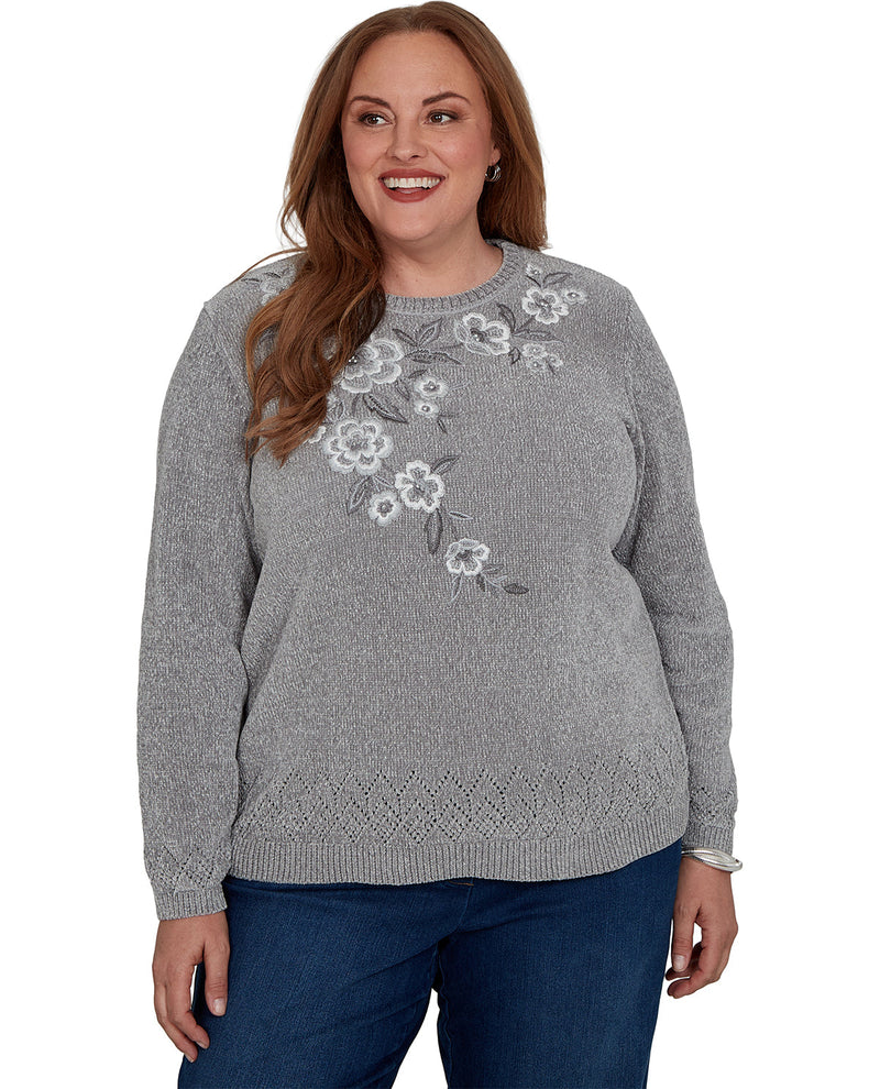 A woman wearing a heather gray knit sweater with white and gray floral embroidery on the chest, paired with dark blue jeans, against a plain white background.