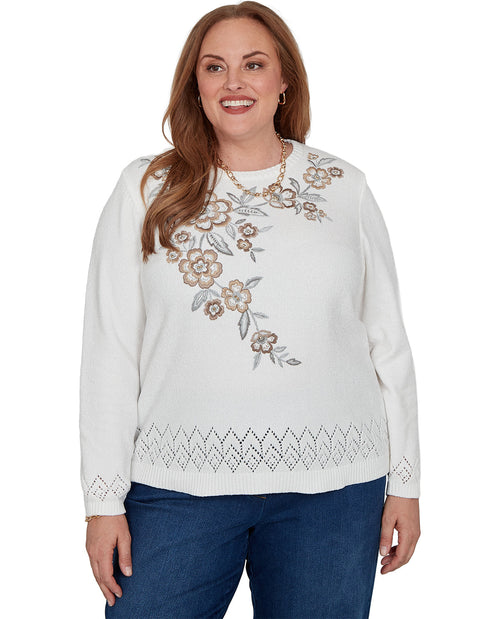 A woman wearing a white long-sleeve knit sweater with floral embroidery on the chest and a geometric lace-like hem, paired with blue jeans against a plain white background.