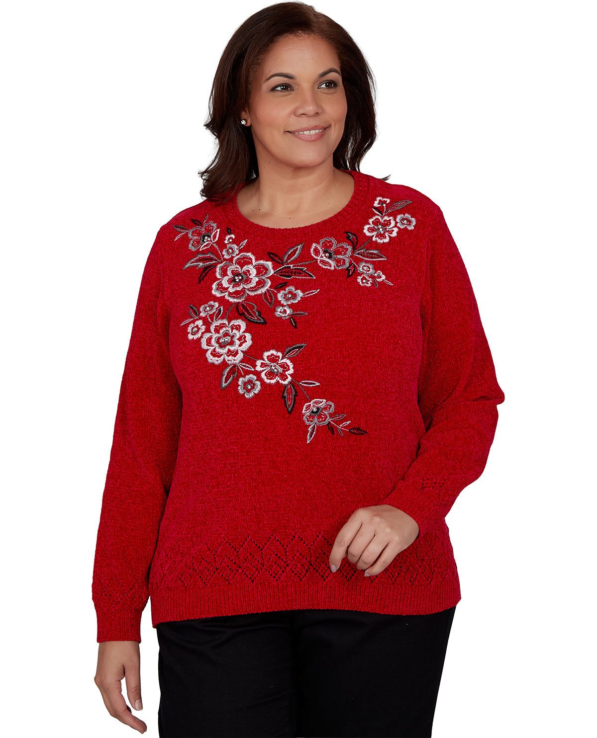A woman wearing a bright red crewneck sweater with a floral embroidered pattern on the chest, paired with black pants, against a plain white background.