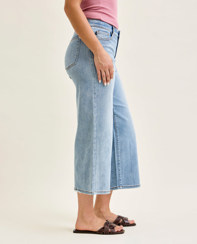A side-profile view of a model wearing light-wash blue cropped flare jeans, a pink top, and dark brown slide sandals against a plain beige studio background.