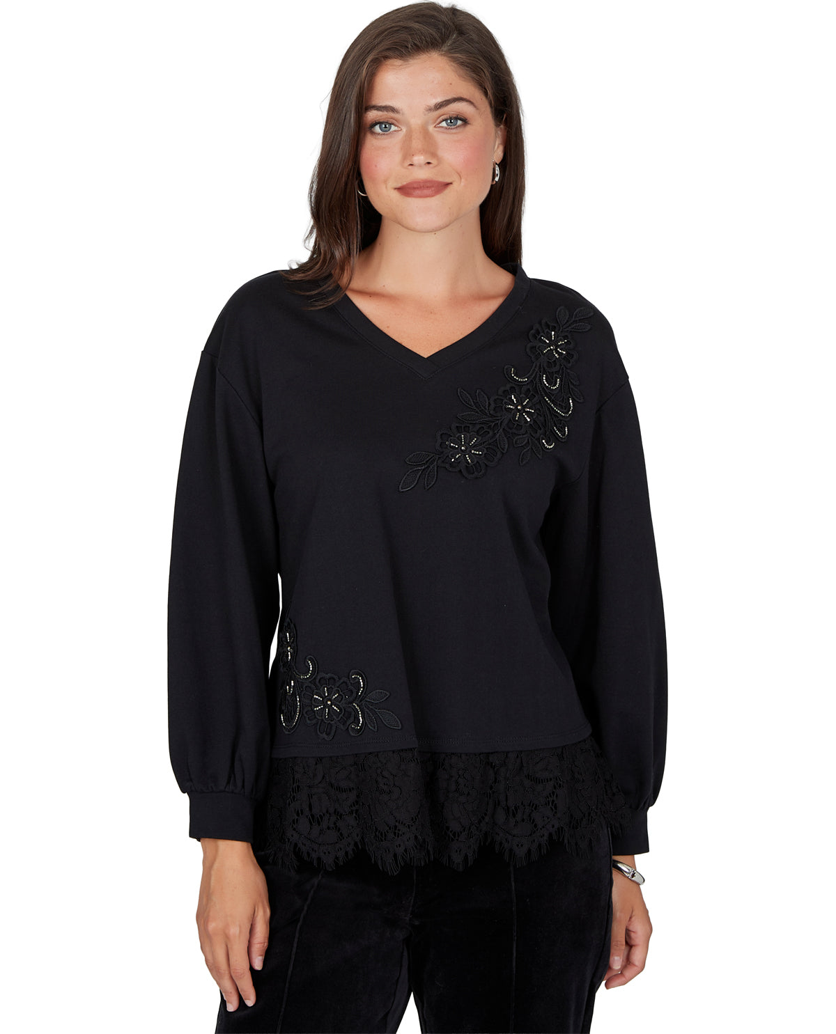 A woman modeling a black long-sleeve V-neck top with floral embroidery on the chest and shoulder, a lace hem, and black pants against a plain white background.
