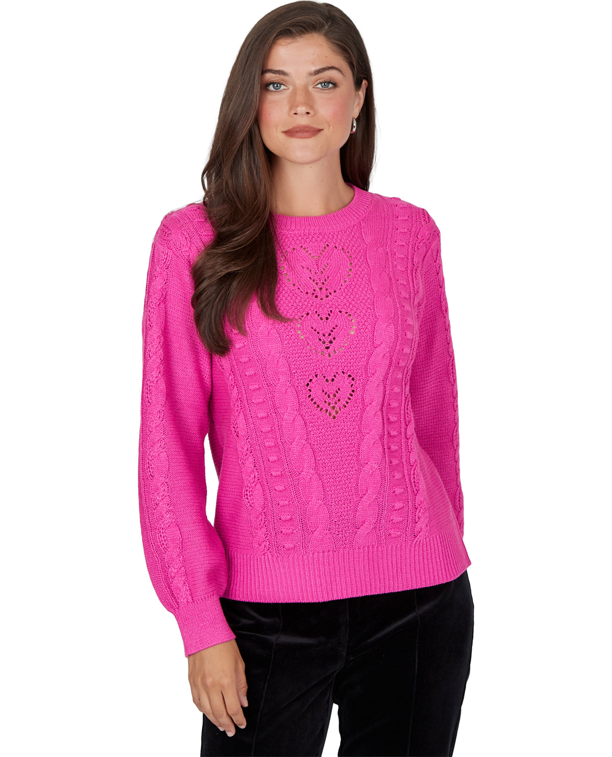 A woman modeling a bright pink cable-knit sweater with heart-shaped cutout details on the front, paired with black pants, against a plain white background.