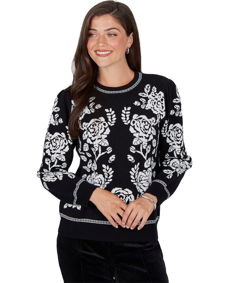 A woman wearing a black crewneck sweater with a white floral pattern and decorative trim, paired with black pants, against a plain white background.
