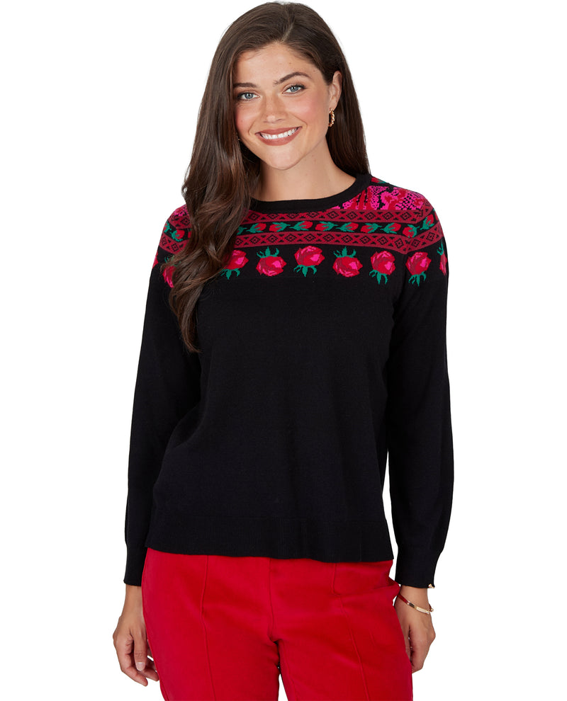 A woman modeling a black knit sweater with a colorful embroidered yoke featuring red roses and green leaves, paired with bright red pants, against a plain white background.