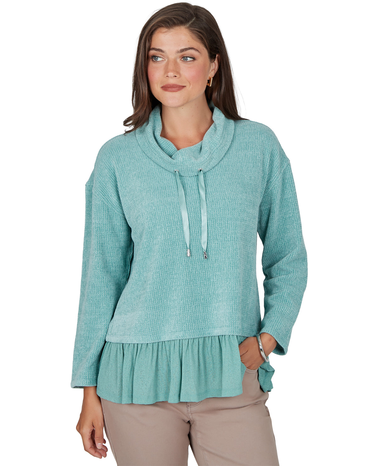 A woman modeling a light teal knit sweater with a cowl neck, drawstring detail, and a ruffled hem, paired with beige pants against a plain white background.