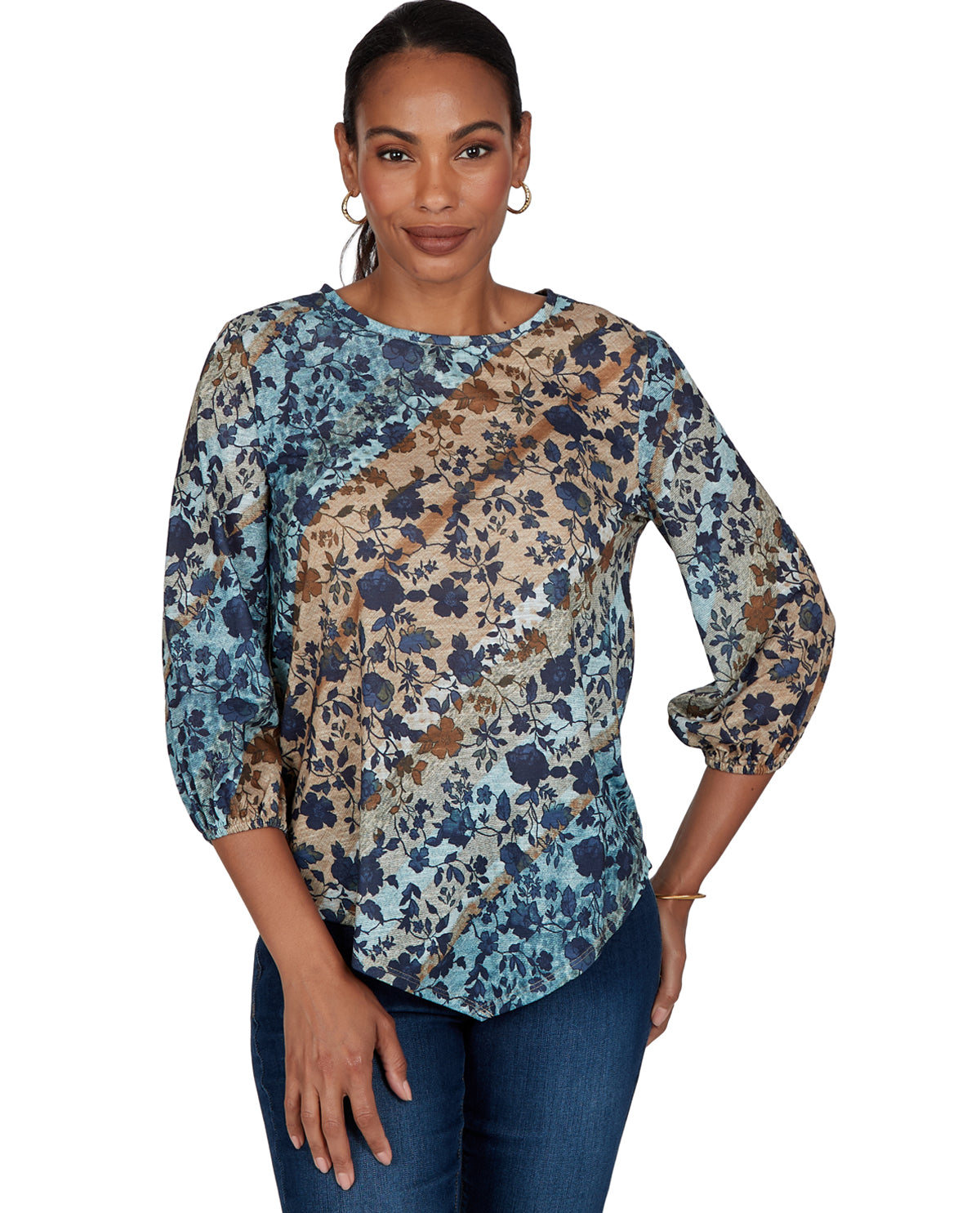A woman modeling a three-quarter sleeve blouse with a diagonal split design in blue, teal, and brown floral print, paired with blue jeans against a plain white background.
