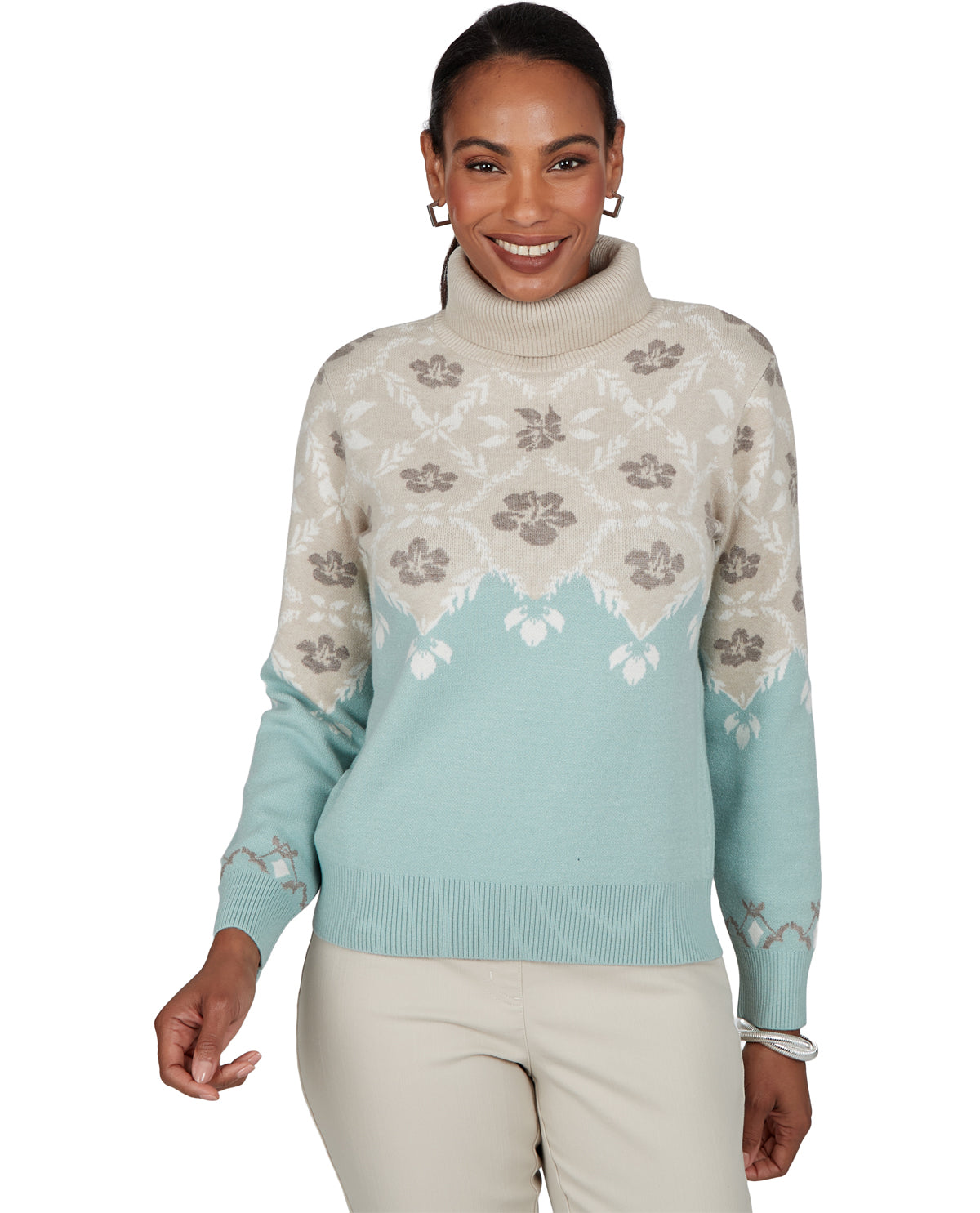 A woman modeling a turtleneck sweater with a cream and light blue color-blocked design and floral patterns, paired with beige pants against a white background.
