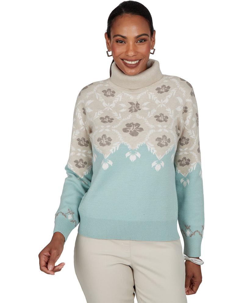 A woman modeling a turtleneck sweater with a cream and light blue color-blocked design and floral patterns, paired with beige pants against a white background.