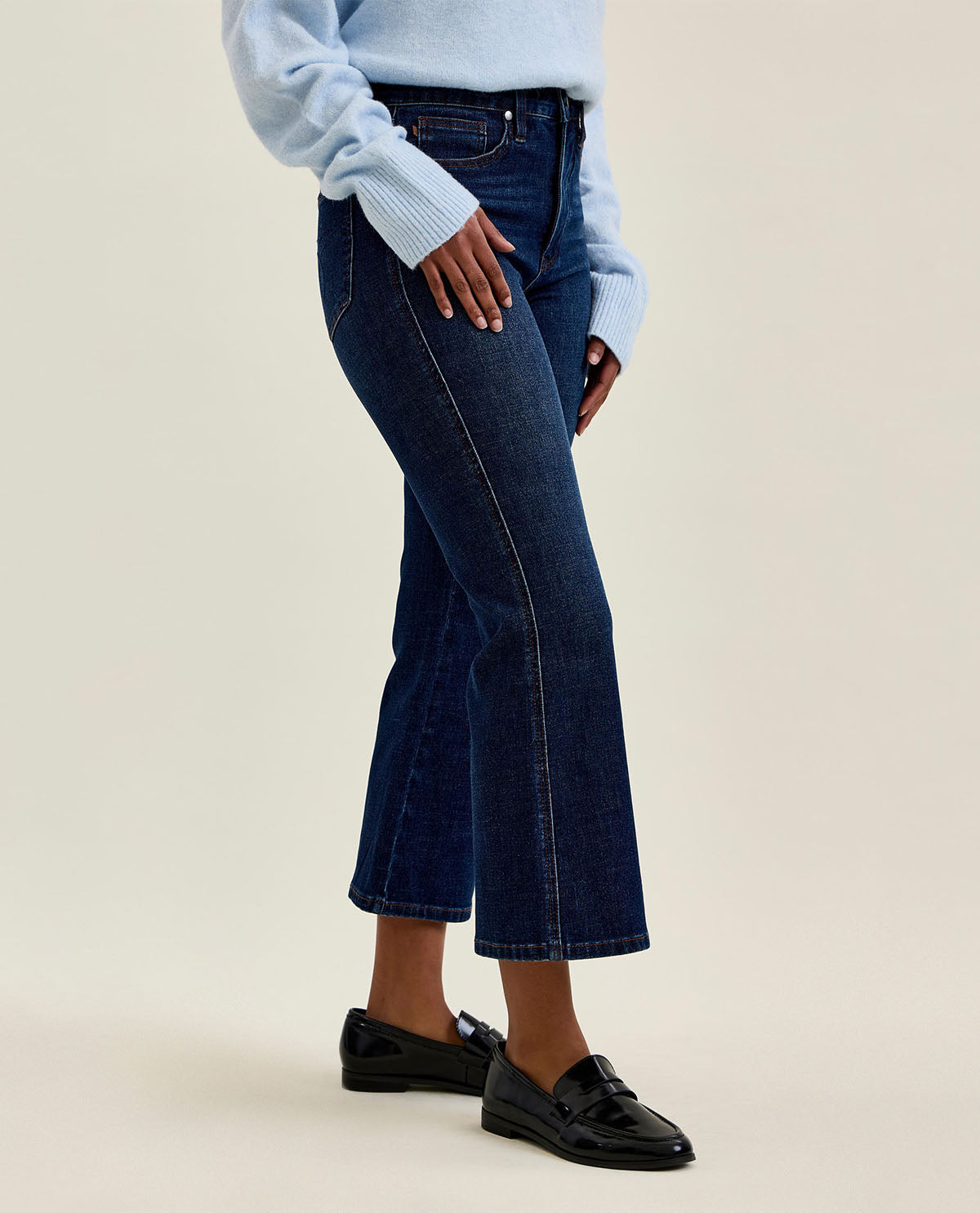 A model wearing dark blue cropped flare jeans, a light blue ribbed sweater, and black loafers, shown from the waist down against a solid beige studio background.