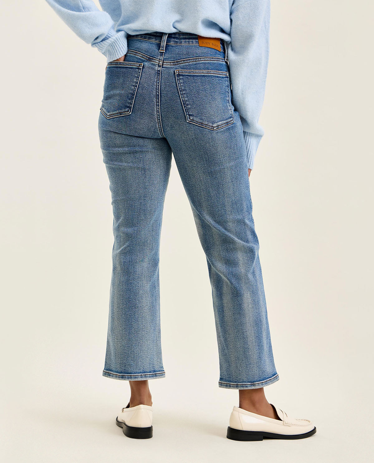 A person shown from the back wearing light blue cropped jeans with a straight-to-medium leg, styled with a light blue long-sleeve top and white loafers against a plain light beige studio background.