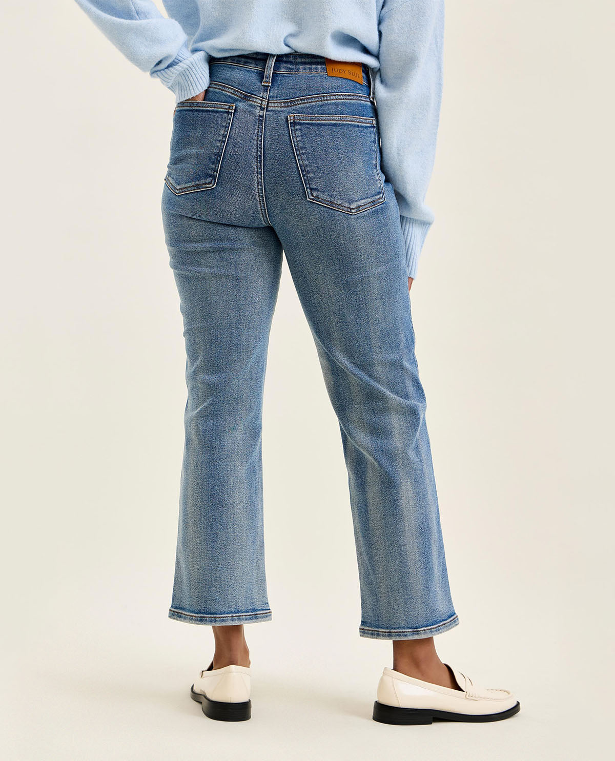 A person shown from the back wearing light blue cropped jeans with a straight-to-medium leg, styled with a light blue long-sleeve top and white loafers against a plain light beige studio background.