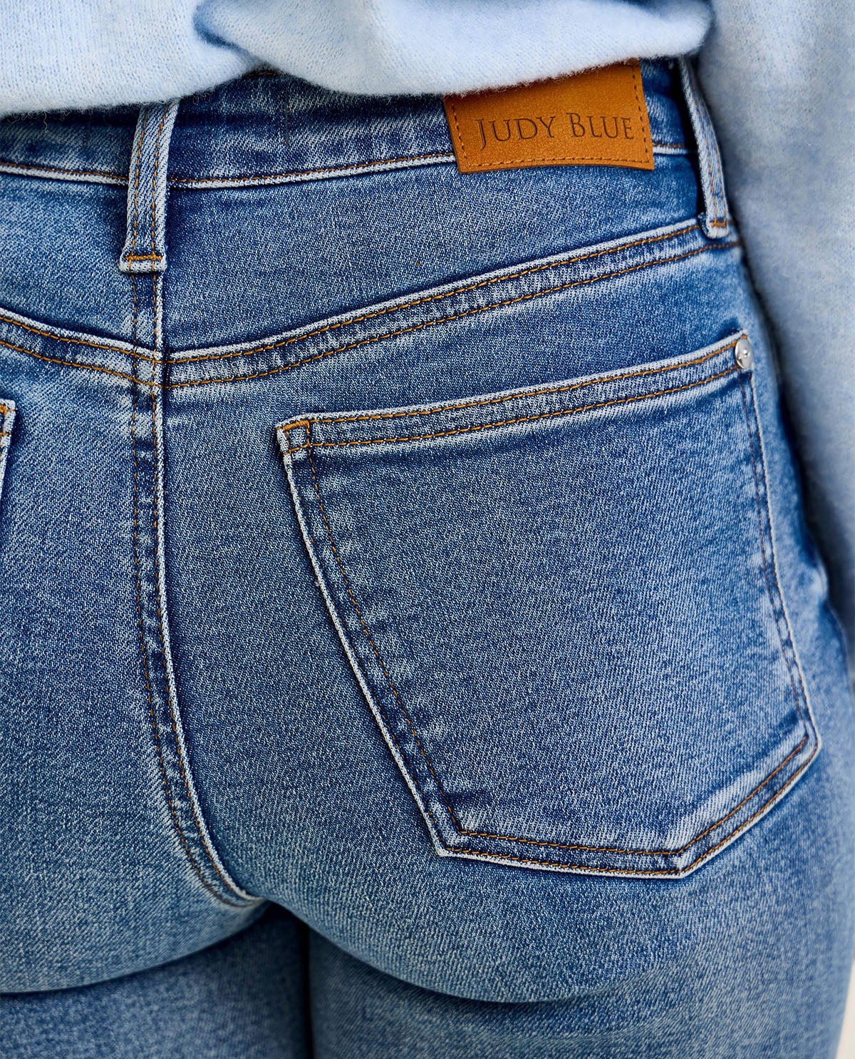 Close-up of the back of light blue denim jeans with a tan leather patch reading 'JUDY BLUE' at the waistband, worn with a light blue top.