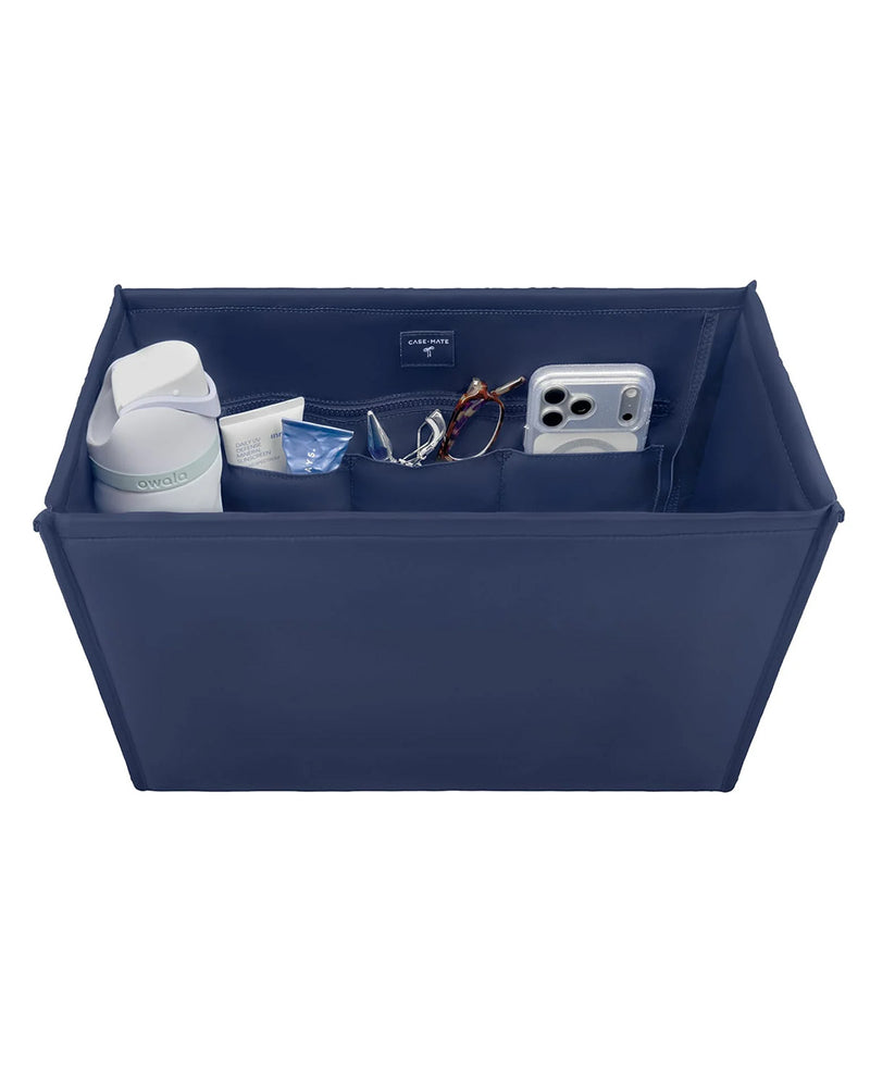 A navy blue fabric organizer tray with multiple compartments, shown holding various personal items like a white bottle, a tube, eyeglasses, and a smartphone. The organizer is photographed on a clean white background.