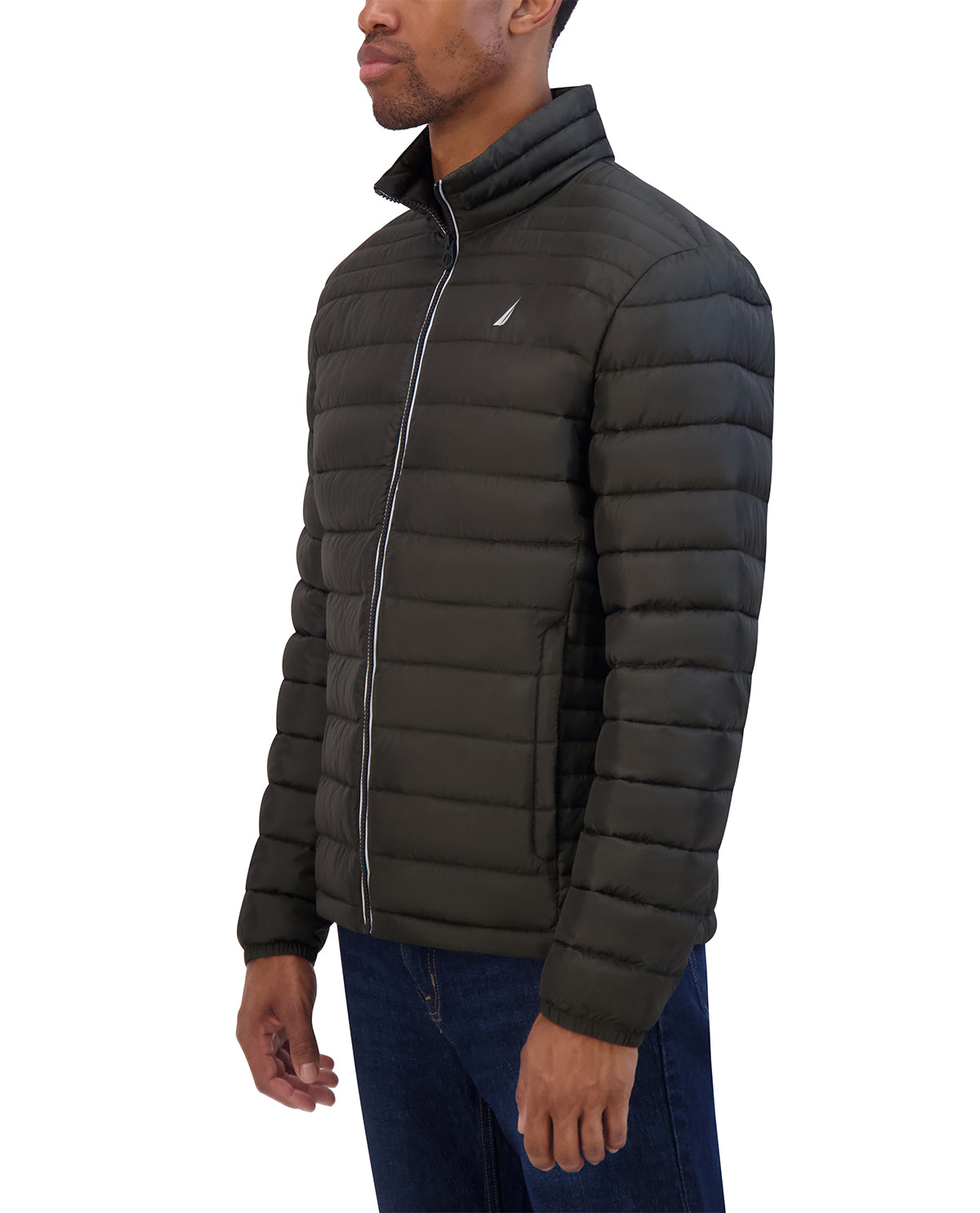 A man wearing a dark olive green puffer jacket with a high collar and silver zipper, paired with blue jeans, shown from a side profile against a white background.