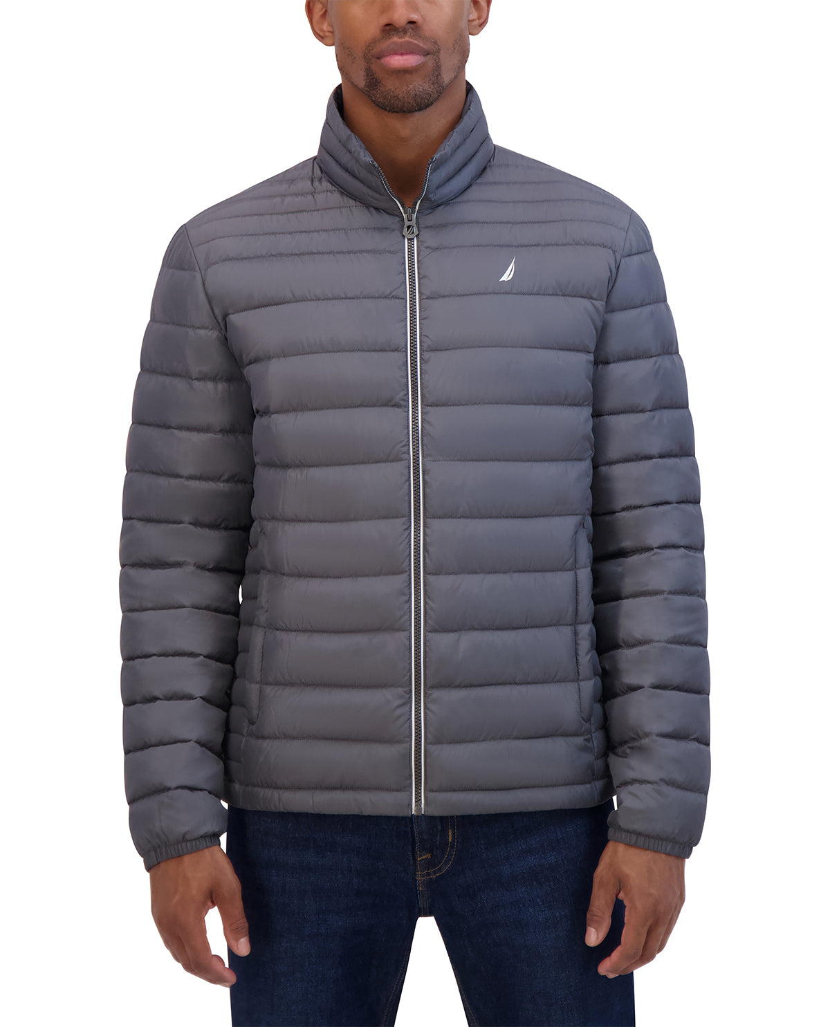 A man wearing a gray horizontally quilted puffer jacket with a stand-up collar and a small white Nautica sail logo on the left chest, paired with dark blue jeans against a plain white background.