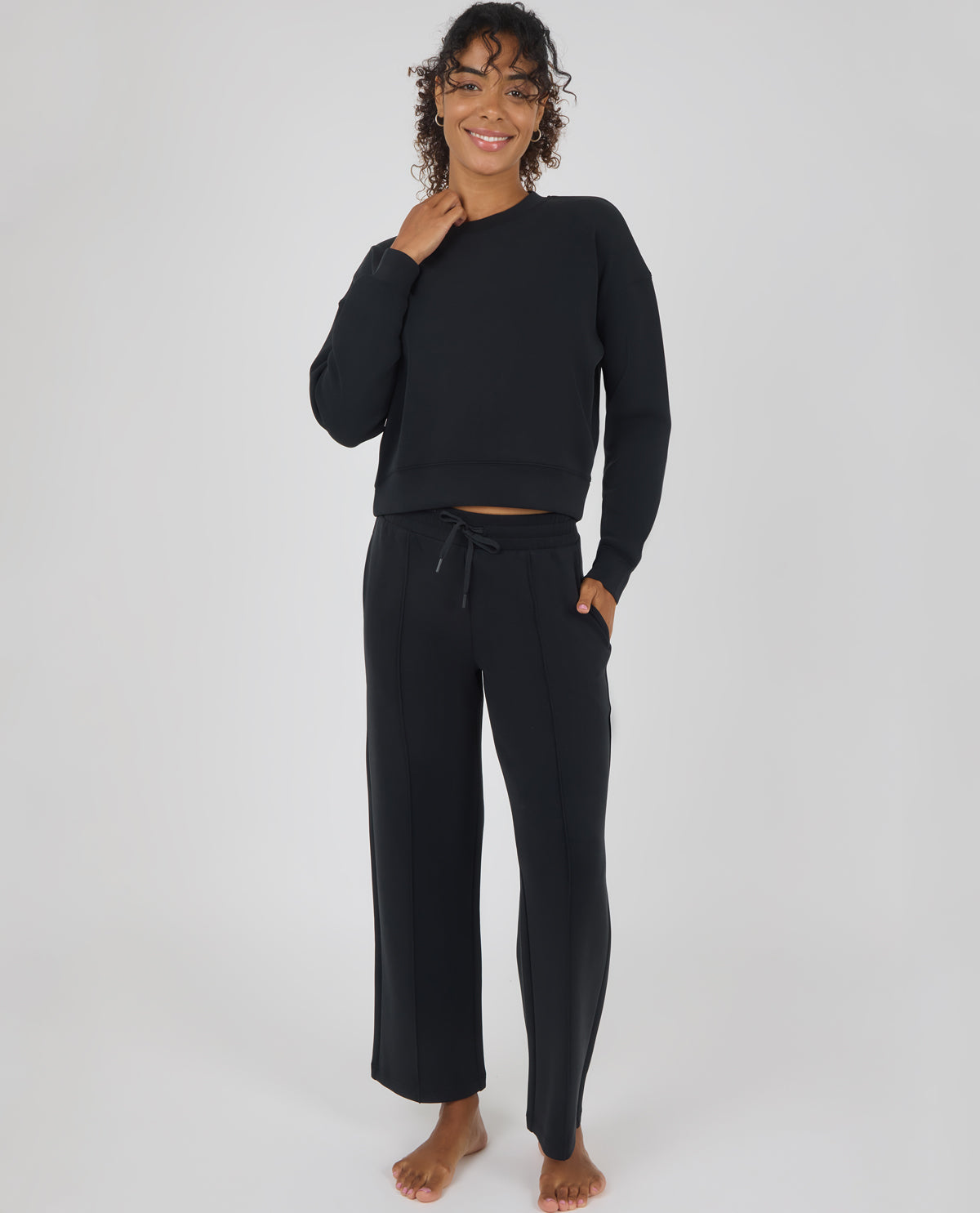 A woman modeling a black two-piece loungewear set consisting of a cropped long-sleeve sweatshirt and matching wide-leg drawstring pants, photographed against a neutral light gray studio background.