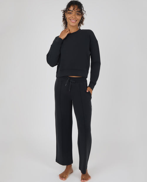 A woman modeling a black two-piece loungewear set consisting of a cropped long-sleeve sweatshirt and matching wide-leg drawstring pants, photographed against a neutral light gray studio background.