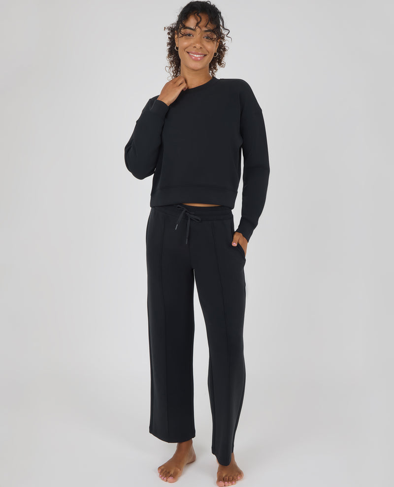 A woman modeling a black two-piece loungewear set consisting of a cropped long-sleeve sweatshirt and matching wide-leg drawstring pants, photographed against a neutral light gray studio background.