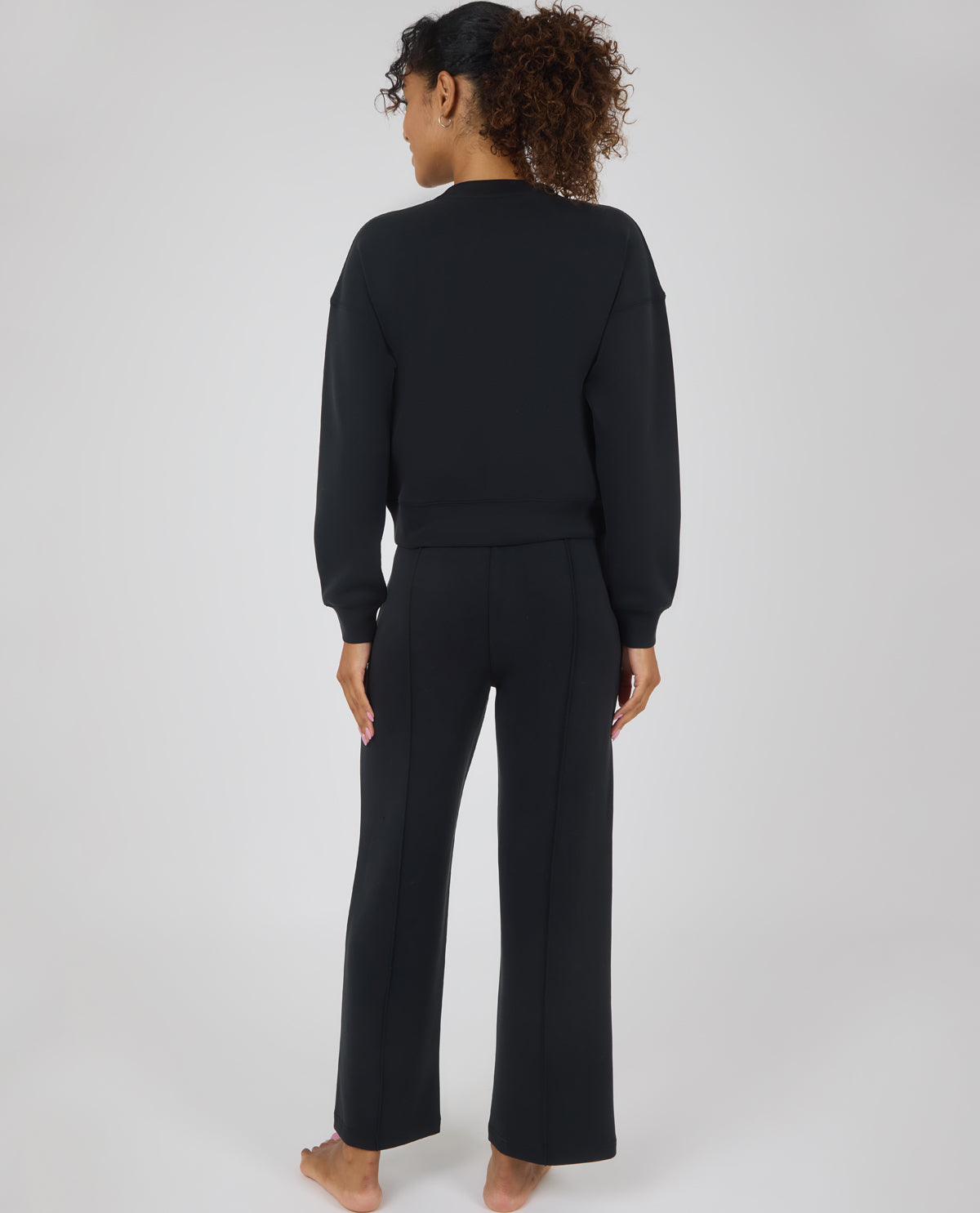 A woman viewed from the back wearing a matching black lounge set consisting of a cropped long-sleeve top and wide-leg pants, standing against a light gray studio background.