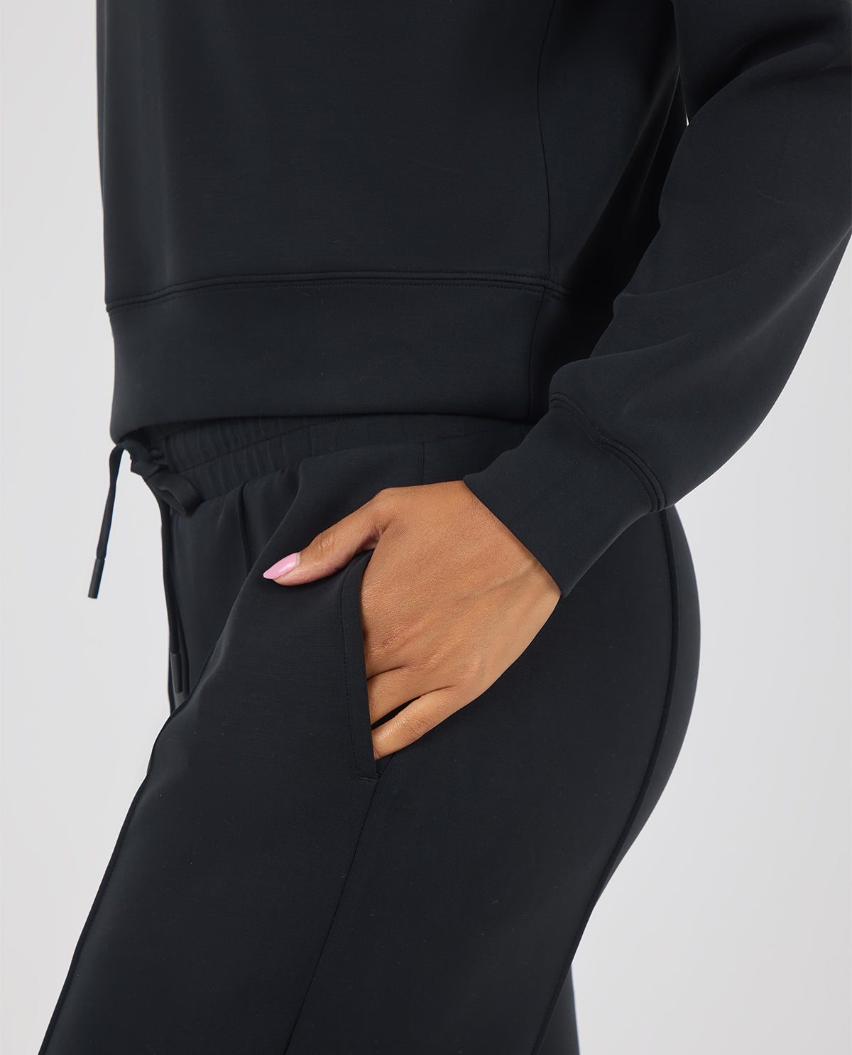Close-up of a person wearing a black sweatshirt and matching black jogger pants, with one hand tucked into the side pocket of the pants against a light gray studio background.