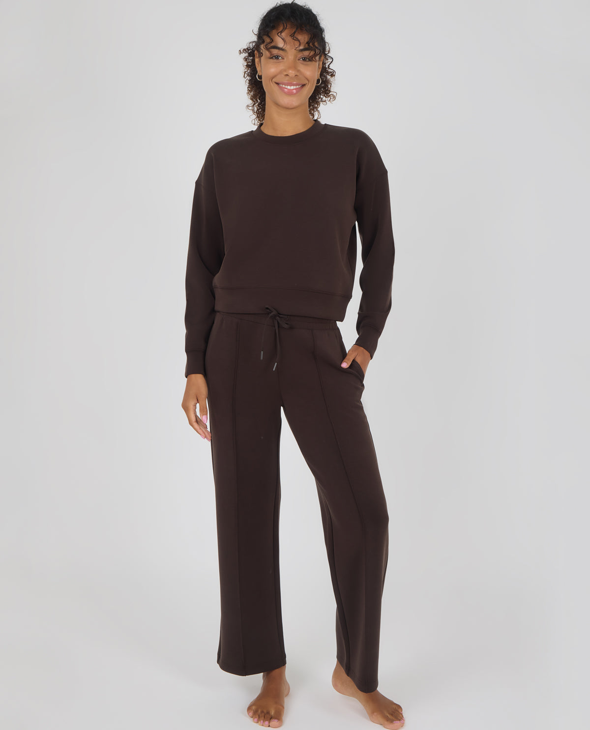 A woman modeling a matching dark brown lounge set consisting of a long-sleeve crewneck sweatshirt and wide-leg drawstring pants, photographed against a neutral light gray studio background.