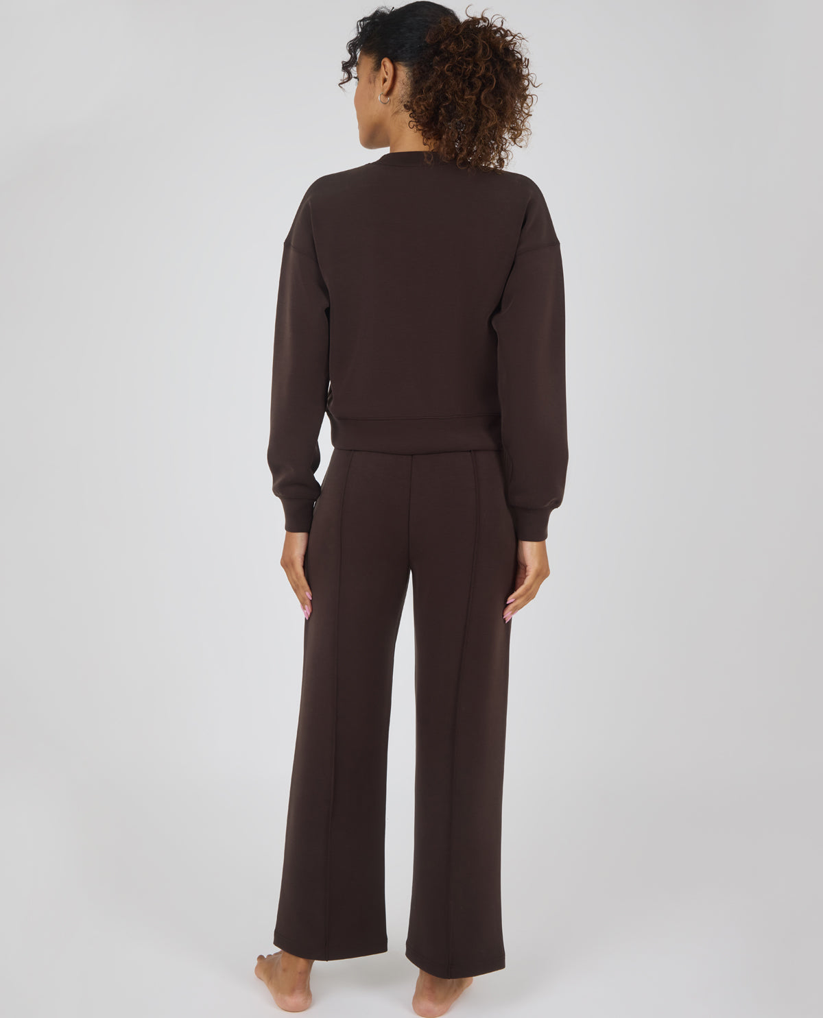 A woman viewed from behind wearing a matching dark brown lounge set consisting of a long-sleeve sweatshirt and wide-leg pants, standing barefoot against a light gray studio background.