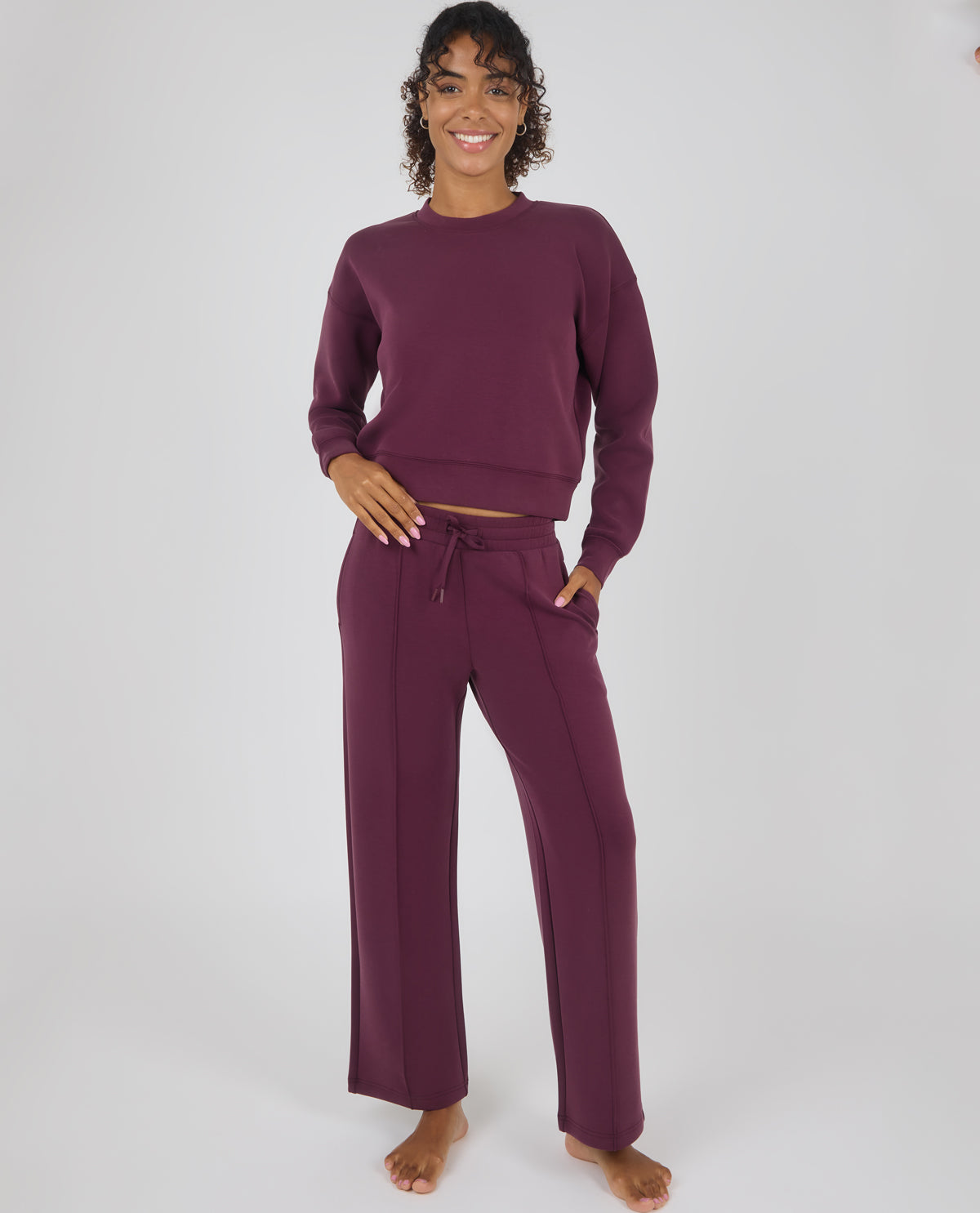 A woman modeling a matching two-piece loungewear set in deep plum purple, featuring a long-sleeve crewneck sweatshirt and wide-leg drawstring pants, against a plain light gray studio background.