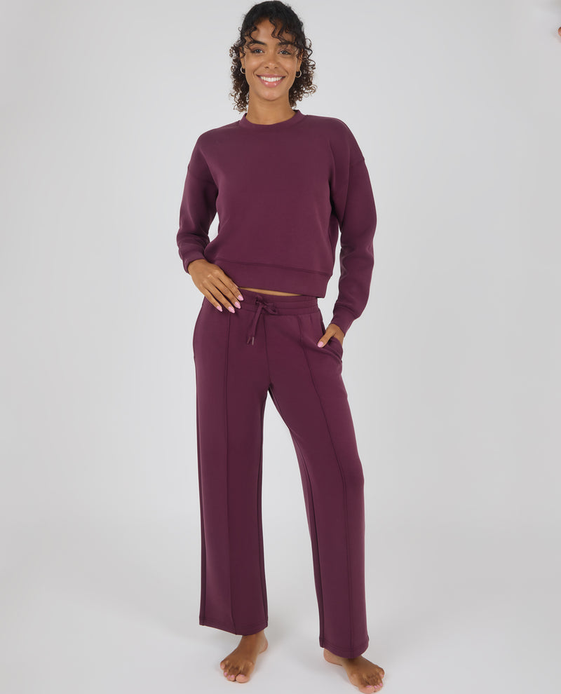 A woman modeling a matching two-piece loungewear set in deep plum purple, featuring a long-sleeve crewneck sweatshirt and wide-leg drawstring pants, against a plain light gray studio background.