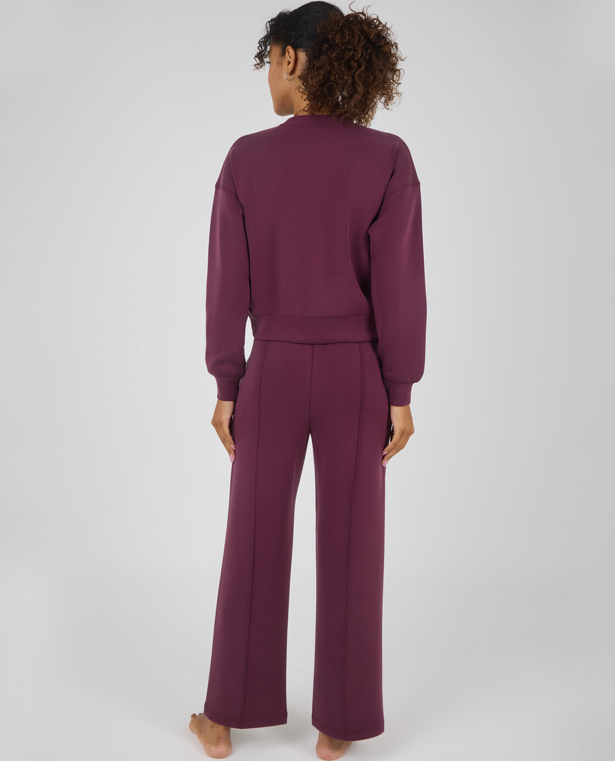 A woman viewed from behind wearing a matching mauve-purple knit lounge set consisting of a long-sleeve cropped sweatshirt and wide-leg pants, against a light gray studio background.