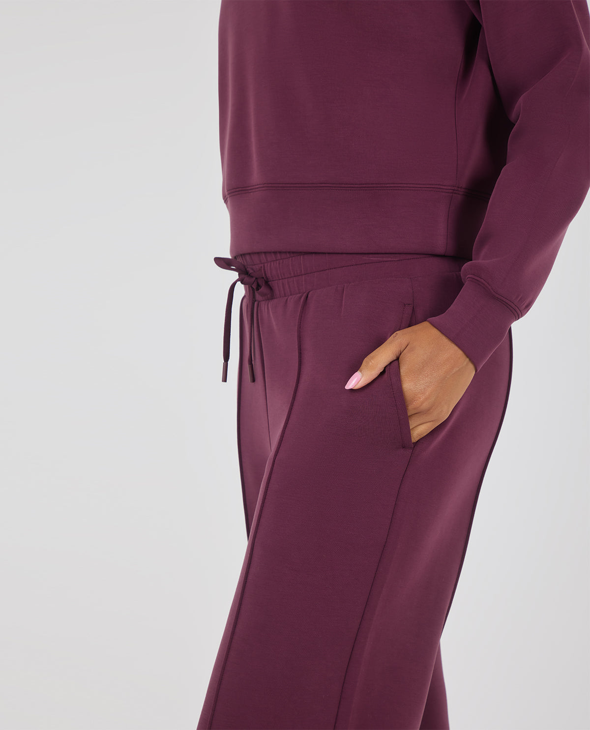 A close-up of a person wearing a matching deep plum sweatshirt and jogger pants set, with one hand in a side pocket against a light gray studio background.