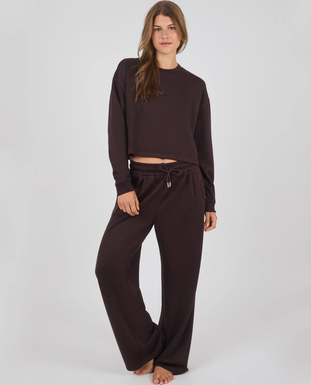 A woman modeling a dark brown two-piece loungewear set consisting of a cropped long-sleeve sweatshirt and matching wide-leg sweatpants, shot against a light gray studio background.