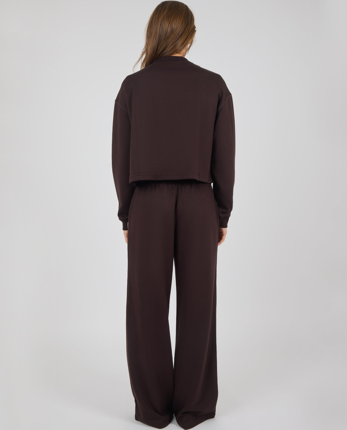 A model viewed from the back wearing a dark brown long-sleeve cropped top and matching wide-leg trousers against a light gray studio background.