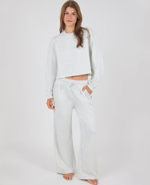 A woman modeling a light heather gray two-piece loungewear set consisting of a cropped long-sleeve sweatshirt and matching wide-leg sweatpants, shot against a seamless light gray studio background.