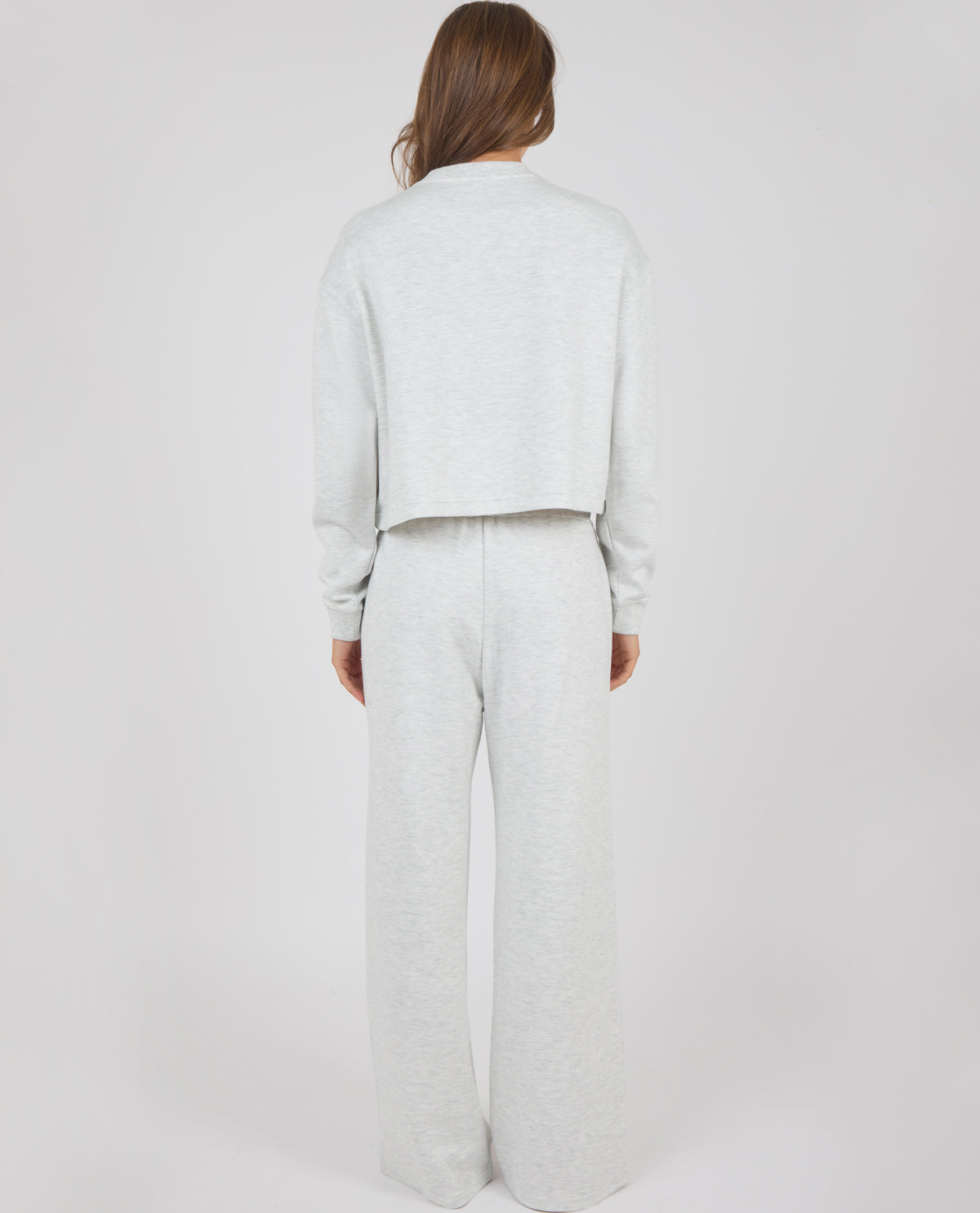 A model viewed from the back wearing a light gray heather cropped sweatshirt and matching wide-leg sweatpants against a plain light gray studio background.