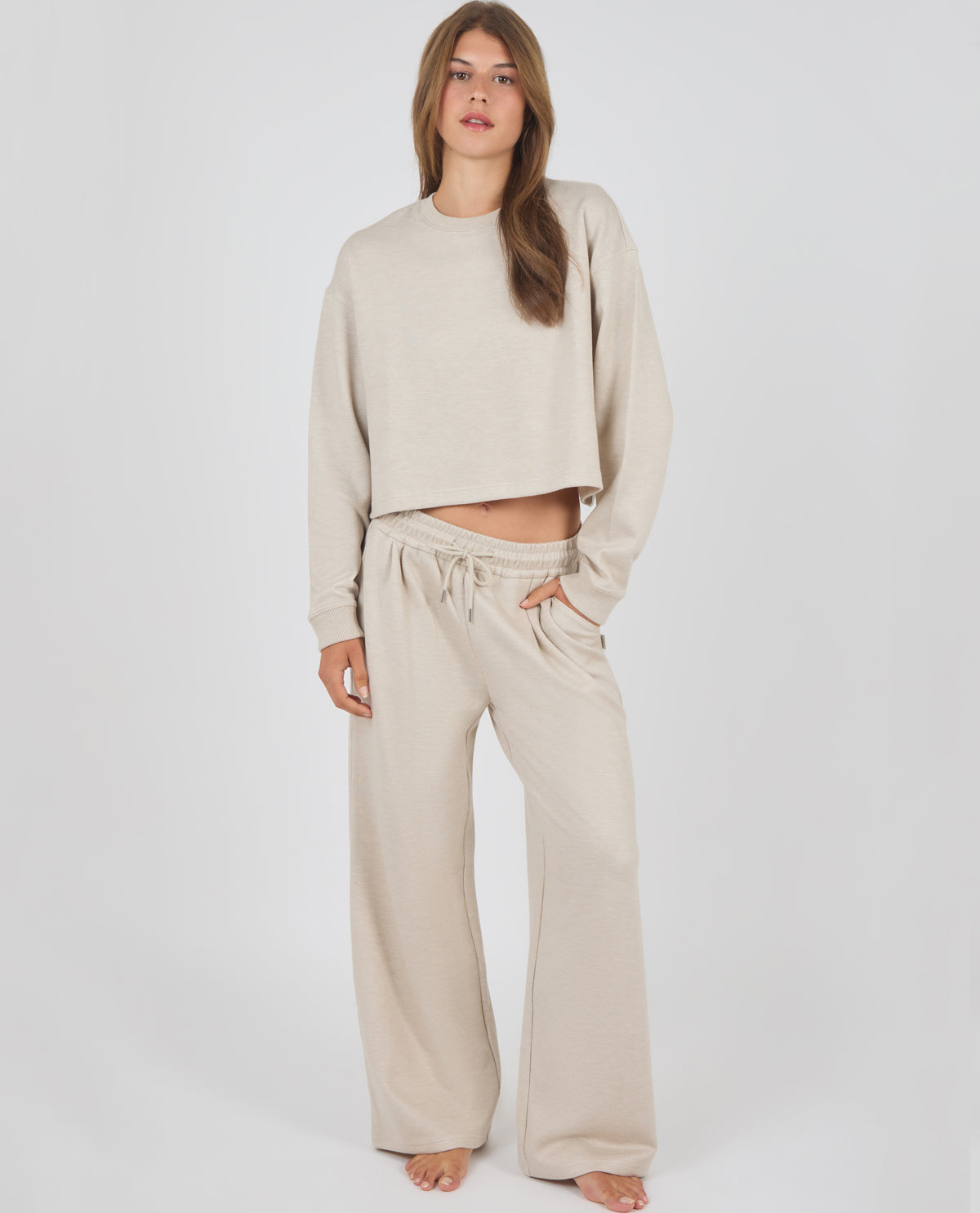 A woman modeling a matching beige loungewear set consisting of a cropped long-sleeve sweatshirt and wide-leg drawstring pants, photographed against a plain light gray studio background.