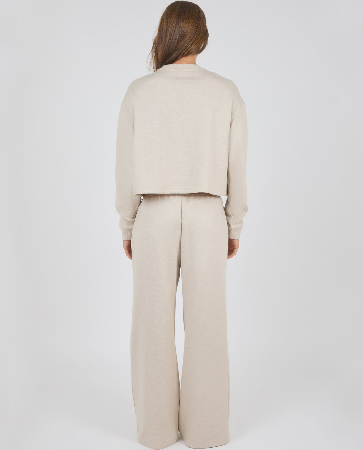 A model viewed from the back wearing a matching beige knit two-piece set consisting of a cropped long-sleeve top and wide-leg trousers, set against a plain light gray background.