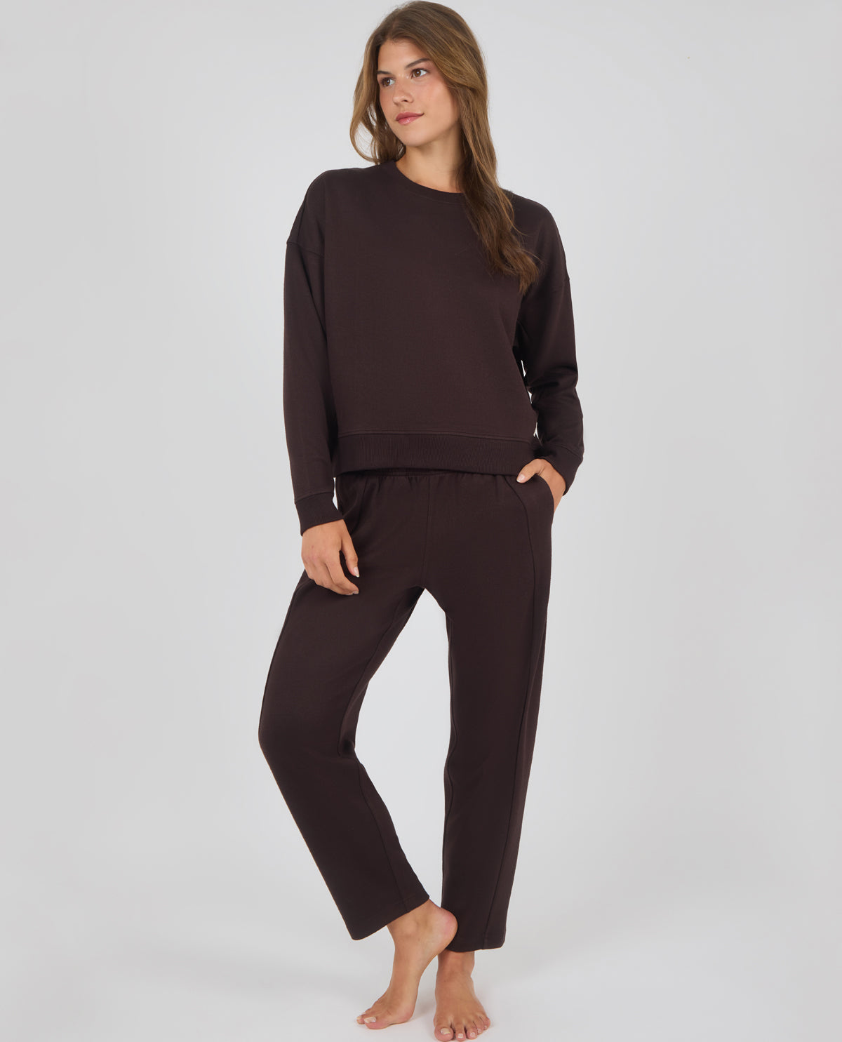 A woman modeling a matching dark brown lounge set consisting of a long-sleeve crewneck sweatshirt and relaxed-fit pants, standing against a light gray studio background.