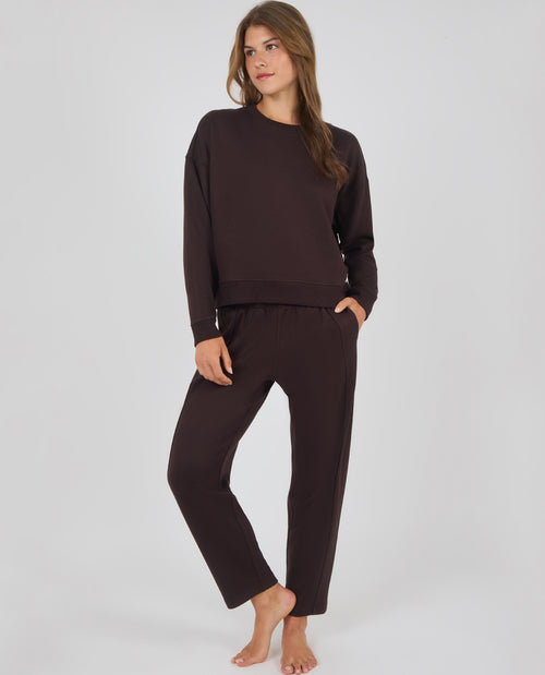 A woman modeling a matching dark brown lounge set consisting of a long-sleeve crewneck sweatshirt and relaxed-fit pants, standing against a light gray studio background.