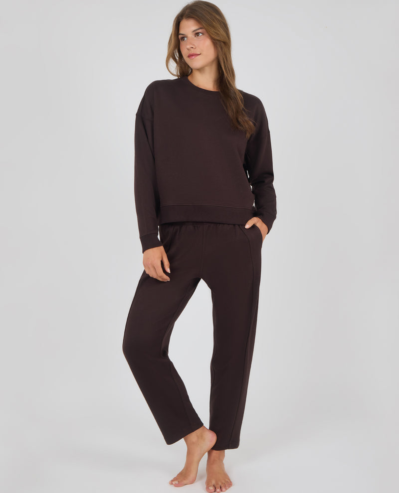 A woman modeling a matching dark brown lounge set consisting of a long-sleeve crewneck sweatshirt and relaxed-fit pants, standing against a light gray studio background.