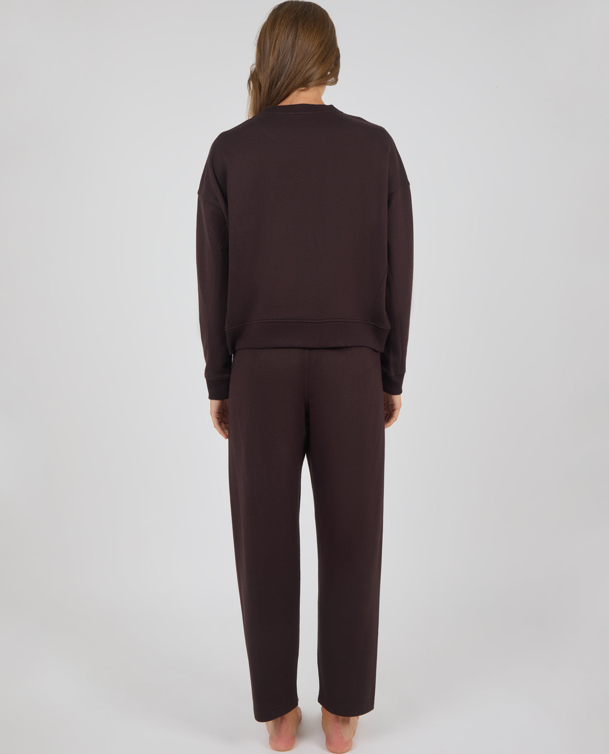 A woman viewed from behind wearing a matching dark brown lounge set consisting of a long-sleeve sweatshirt and wide-leg sweatpants, standing against a light gray studio background.