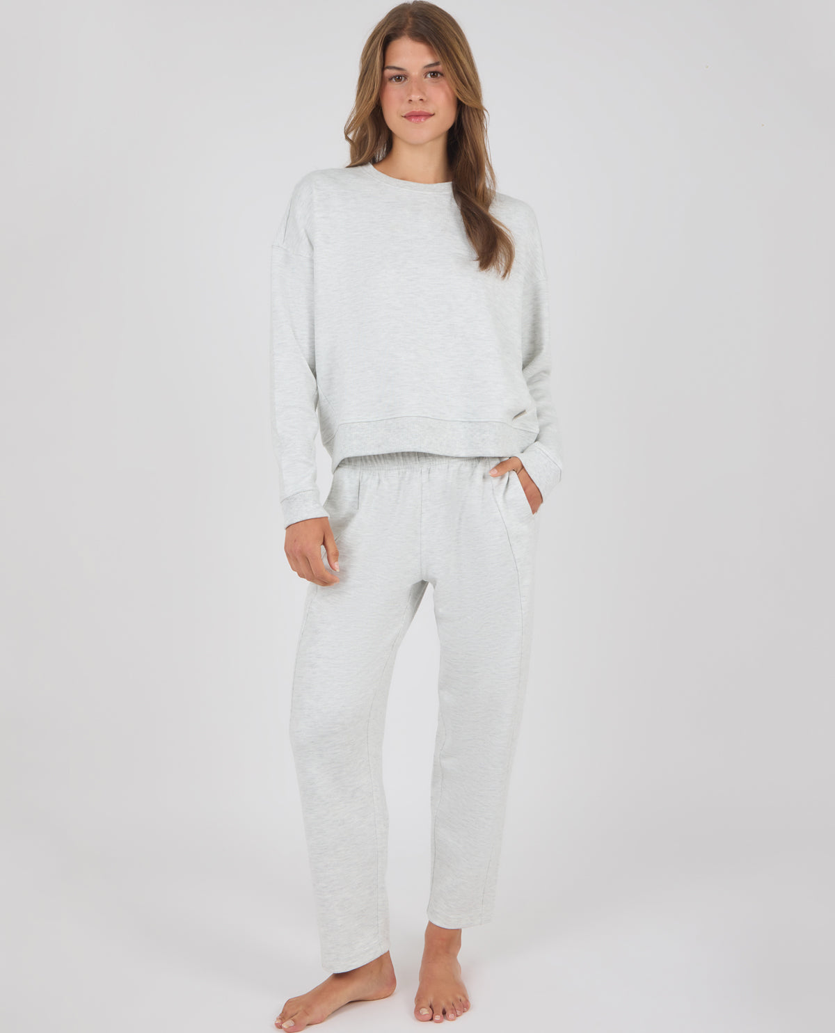 A woman modeling a light gray heathered lounge set consisting of a long-sleeve crewneck sweatshirt and matching jogger pants, standing against a plain light gray studio background.